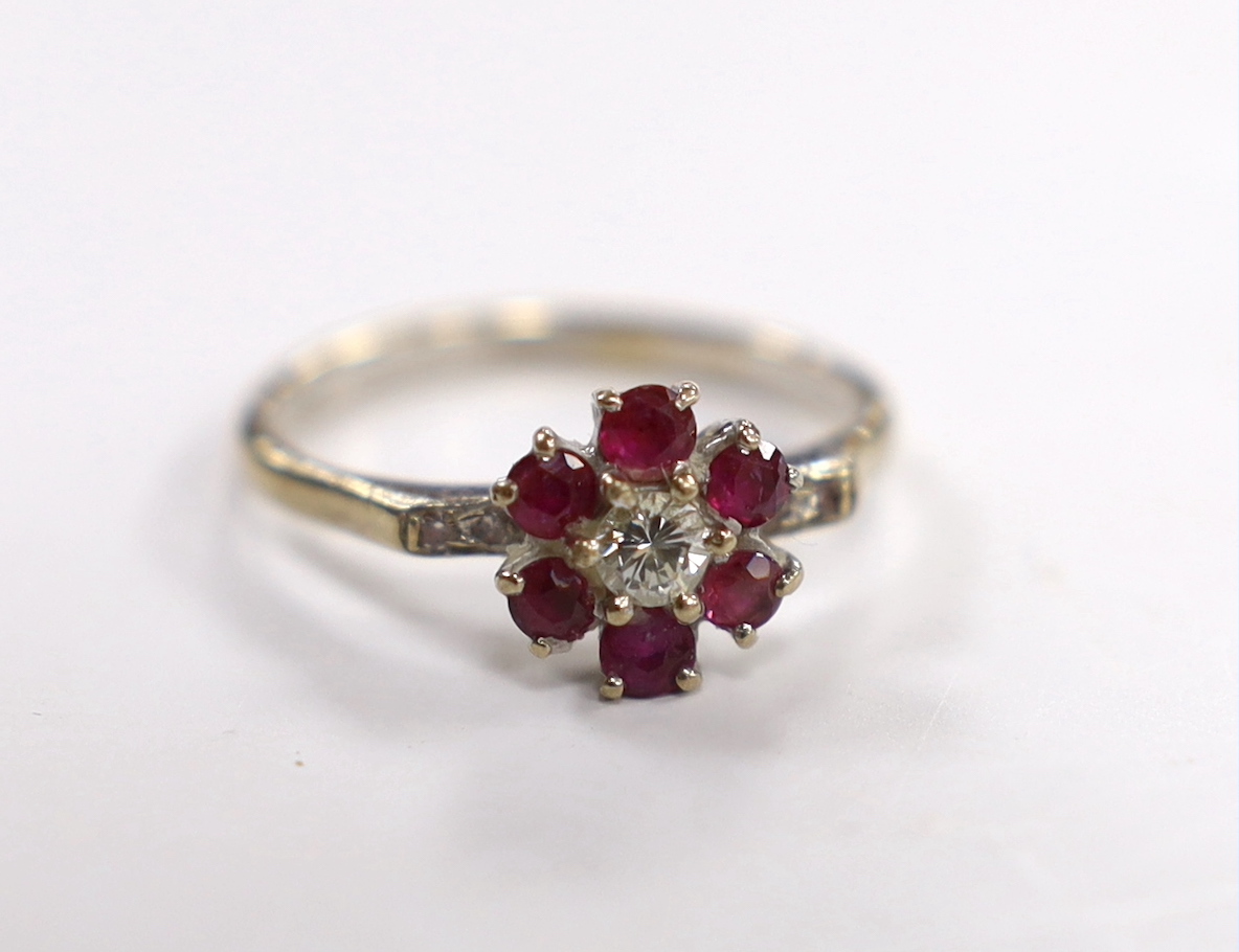 An 18ct white metal, ruby and diamond cluster set flower head ring, with diamond chip set shoulders, size I, gross weight 3.1 grams.